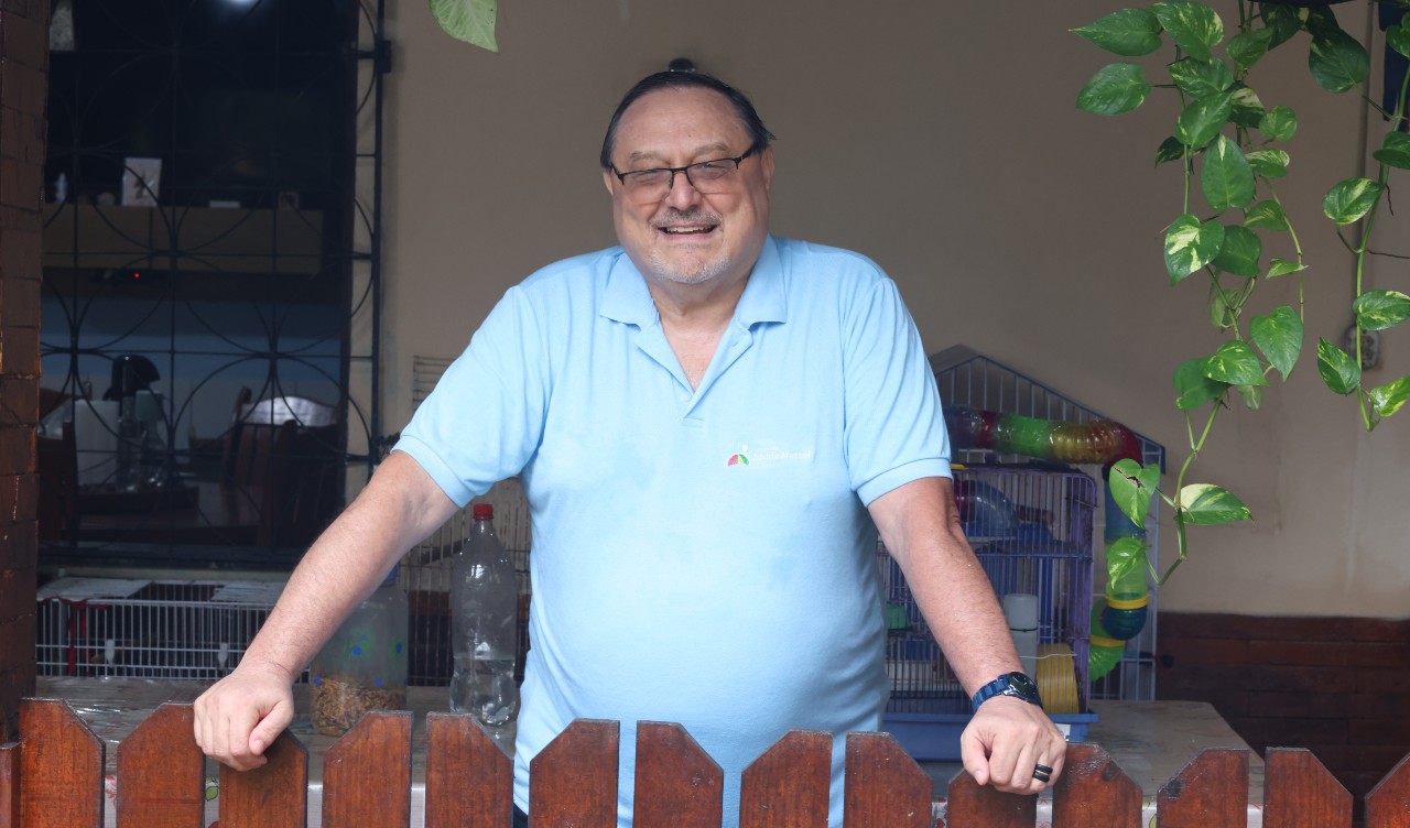 padre rino sorri para a foto atrás de uma cerca em sua casa, ele veste uma camisa polo azul